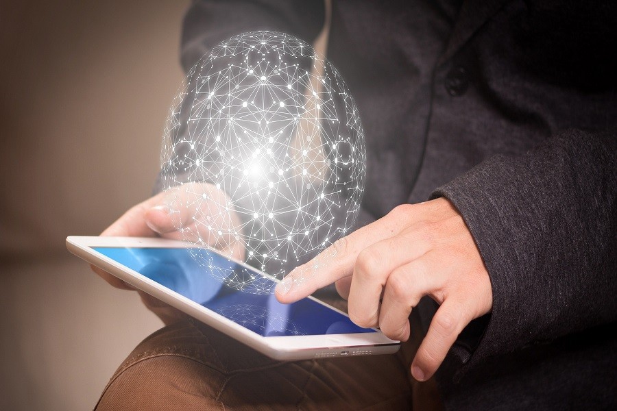 Hands touching a tablet computer as a white graphic sphere of computer topology and interconnected nodes hover above the screen.