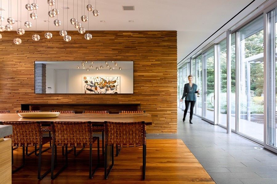 Woman with wine bottle walking into an open concept dining room with sophisticated lighting and a wall of windows.