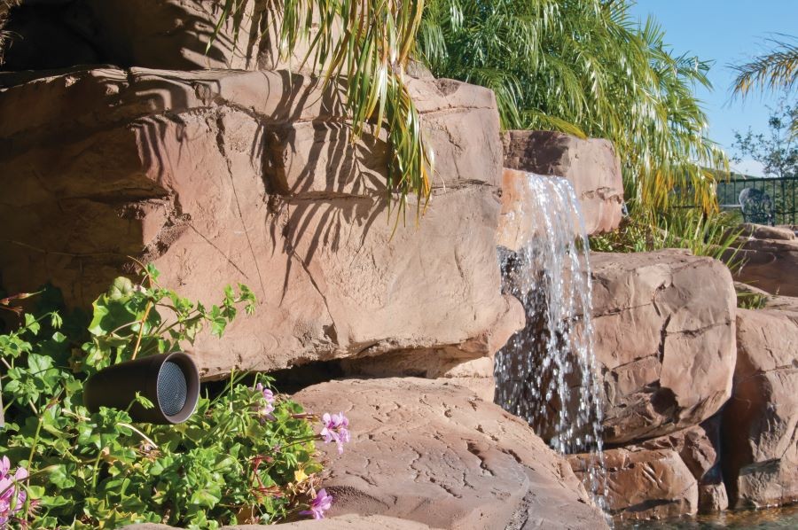 An outdoor speaker next to rocks in a backyard with a small waterfall.